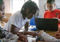 Four 6th grade students collaborate on a hands-on QUEST challenge using a game board and laptop.